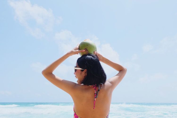 Woman holding coconut while standing at beach against sky