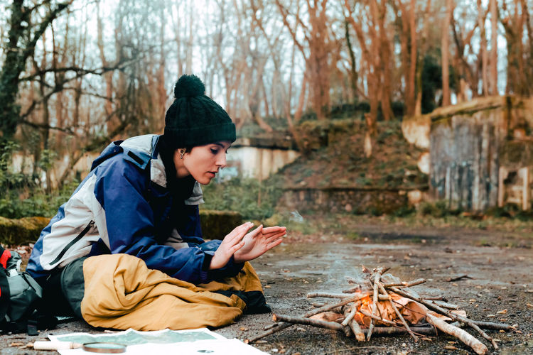 Side view of young female camper in warm clothes warming hands at bonfire while sitting on ground in countryside in autumn day