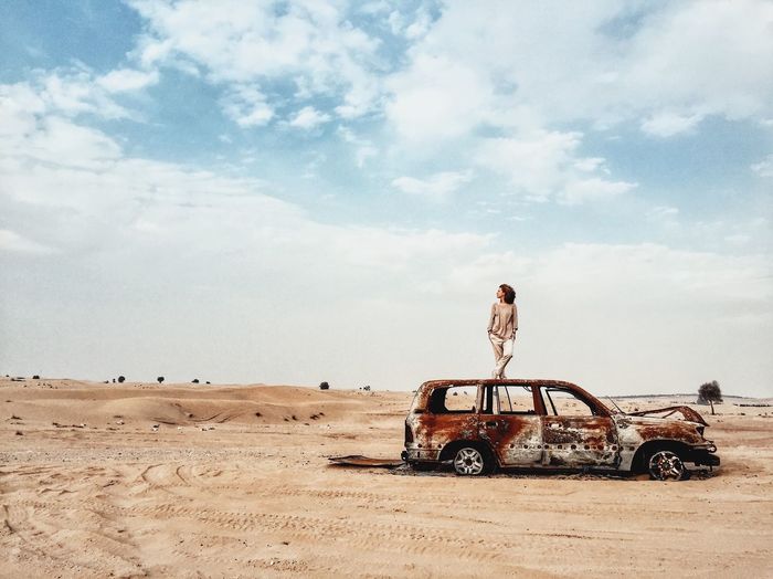 A woman standing on a wreck of the car in the middle of the desert.