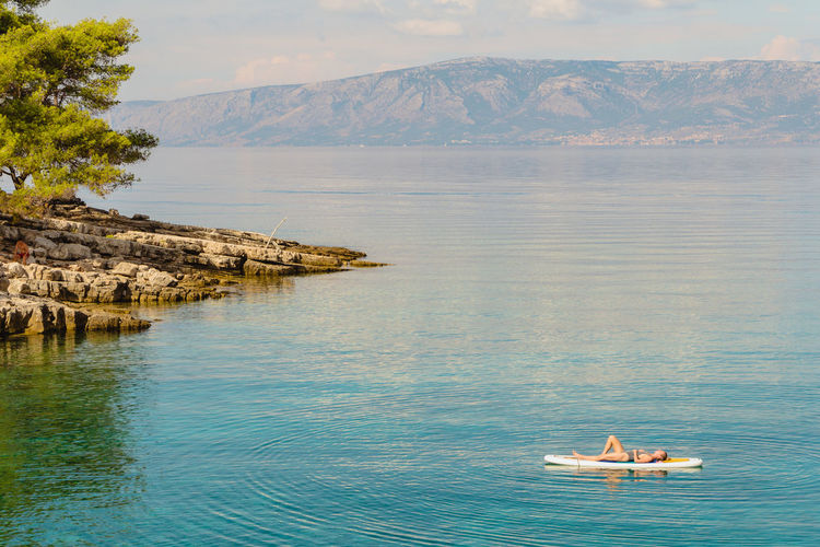 Young woman lays on a paddleboard in a blue bay near a jagged coastline with distant mountains.