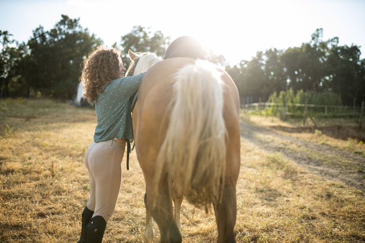 Side view of middle aged female with curly hair putting saddle on back of mare on farm in sunlight