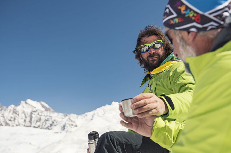 Hikers having drinks on snow covered mountain against clear blue sky during sunny day