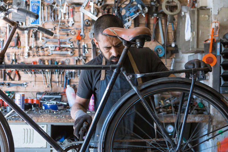 Concentrated male mechanic with beard and tattoos in gloves repairing bicycle in modern workshop