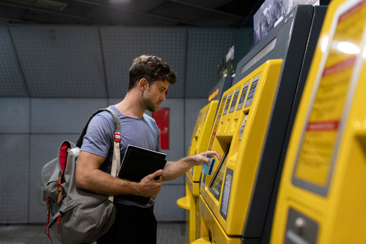 Handsome young male commuter holding book while using ticket machine at station