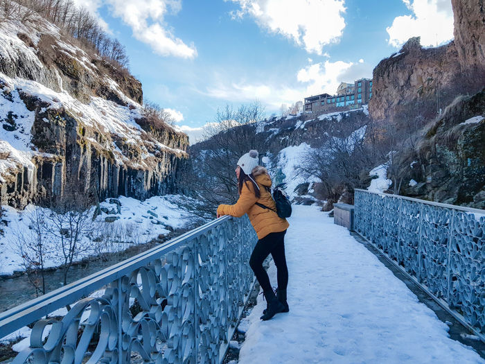 Side view of young woman standing on snow covered footbridge against sky