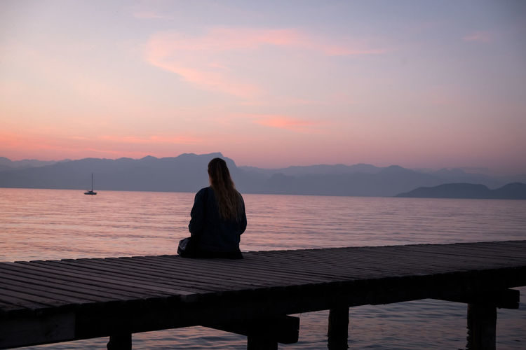 Woman sitting on pier at sea against sky during sunset