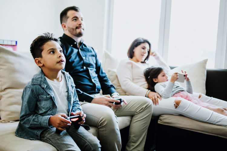 Concentrated young ethnic father with son playing video game using game pads while sitting on comfortable sofa near mother and daughter browsing smartphone