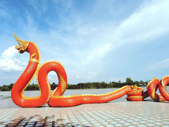Orange snake sculpture at riverbank against sky