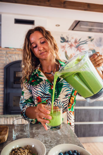 Attractive female pouring healthy green smoothie from blender bowl into glass while standing near table in light kitchen at home