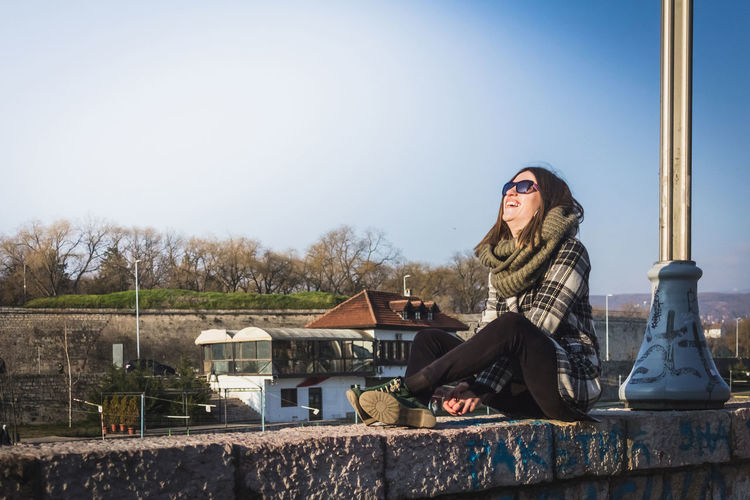 Young woman laughing while sitting on retaining wall against sky