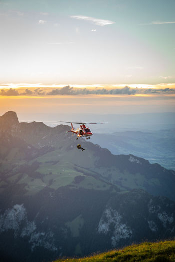 Helicopter flying over mountains against sky during sunset