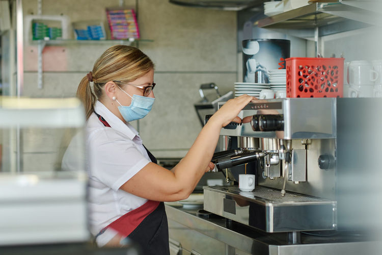 Side view of female barista in medical mask using coffee machine and filling cup with hot espresso while working in cafeteria during coronavirus pandemic