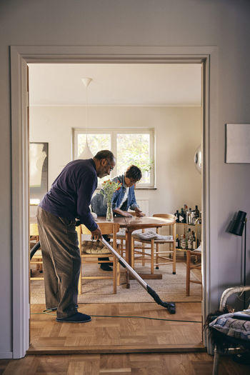 Side view of senior man cleaning floor with vacuum cleaner seen through doorway at home