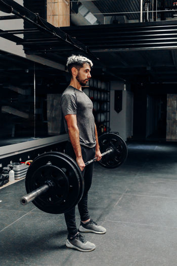 Full body side view of concentrated young bearded male athlete lifting heavy barbell during intense workout in contemporary sports club