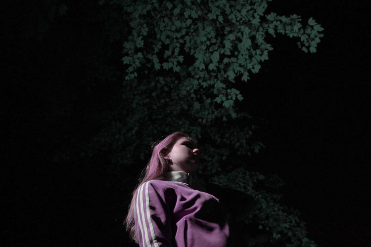Side view of young woman looking away against trees at night