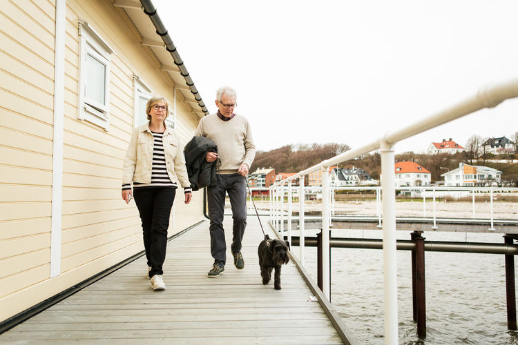 Full length of senior couple walking with schnauzer on pier against sky