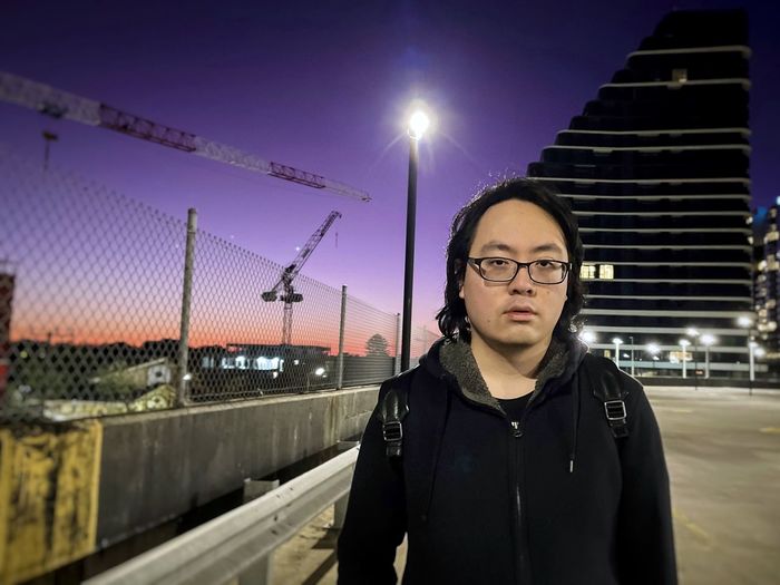 Portrait of young asian man standing on rooftop parking lot against lights and buildings.