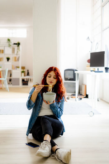 Businesswoman eating noodles with chopsticks while sitting on floor at office