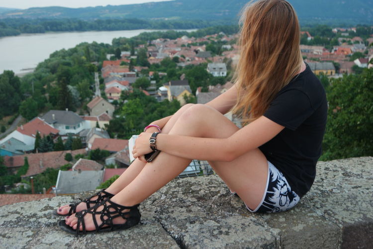 Woman hugging knees while sitting on retaining wall against townscape