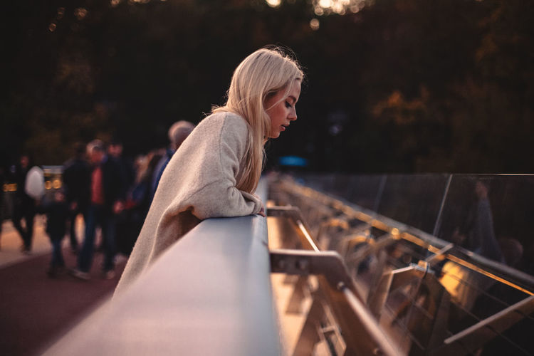 Young woman looking down while standing on bridge in city at sunset