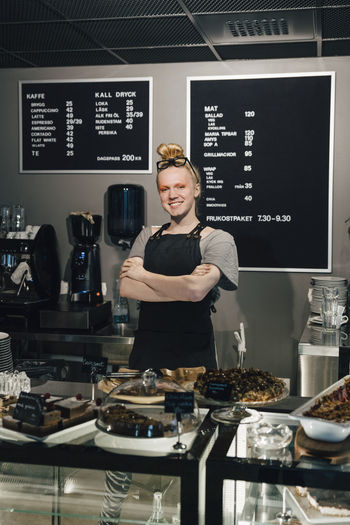 Portrait of smiling barista standing with arms crossed by food variation on counter in illuminated cafeteria