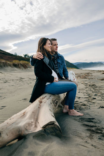 Couple sitting on fallen tree at beach against sky