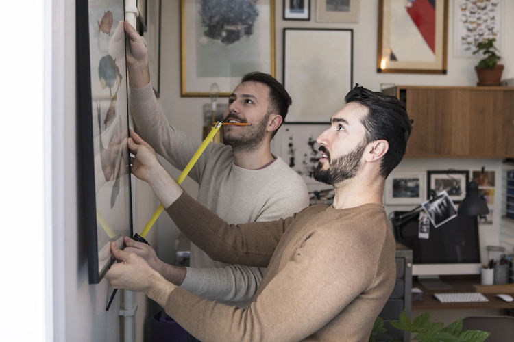 Young man with tape measure assisting partner while hanging painting on wall