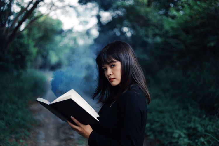 Portrait of woman holding book while standing in forest