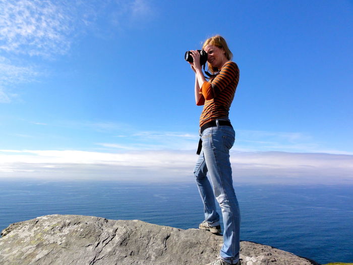 Full length of woman photographing standing on cliff by sea against sky