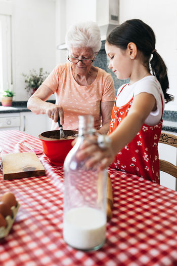 Grandmother in eyewear with whisk near granddaughter with bottle of milk cooking dough at table in house