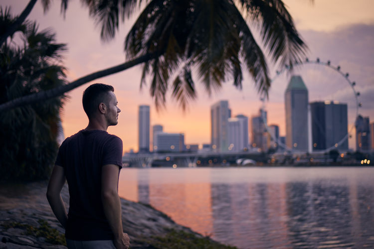 Handsome man standing under palm tree on waterfront of bay and looking at city.