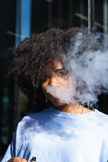 Modern relaxed african american female with curly hair exhaling fume through nose while smoking on street looking at camera