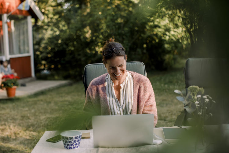 Smiling woman using laptop while sitting at dining table in yard during summer