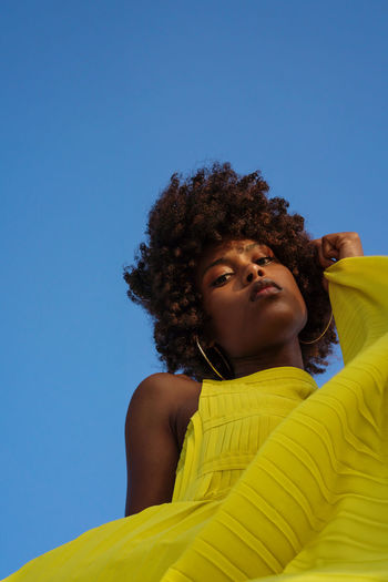 Low angle shot beautiful black woman with afro wearing yellow dress against blue sky