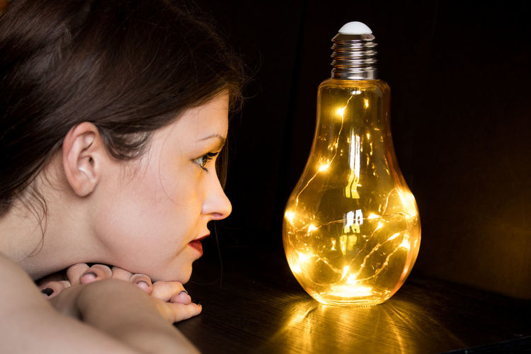 Close-up of young woman looking at illuminated light bulb against black background