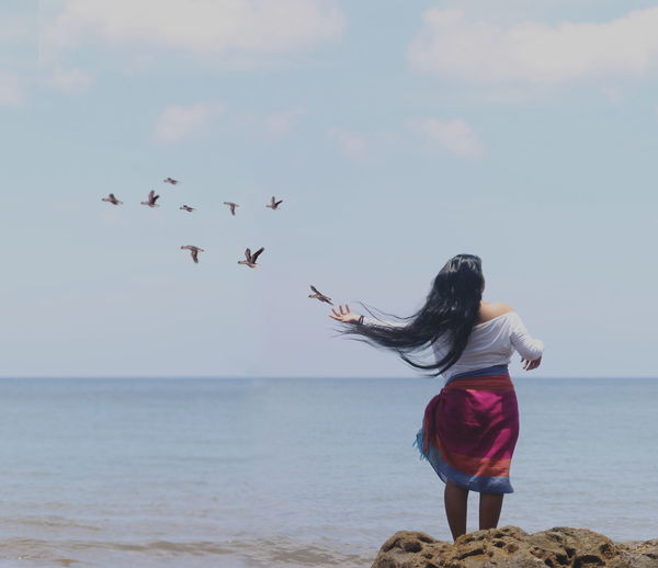Rear view of woman with long hair standing on rock by sea against sky