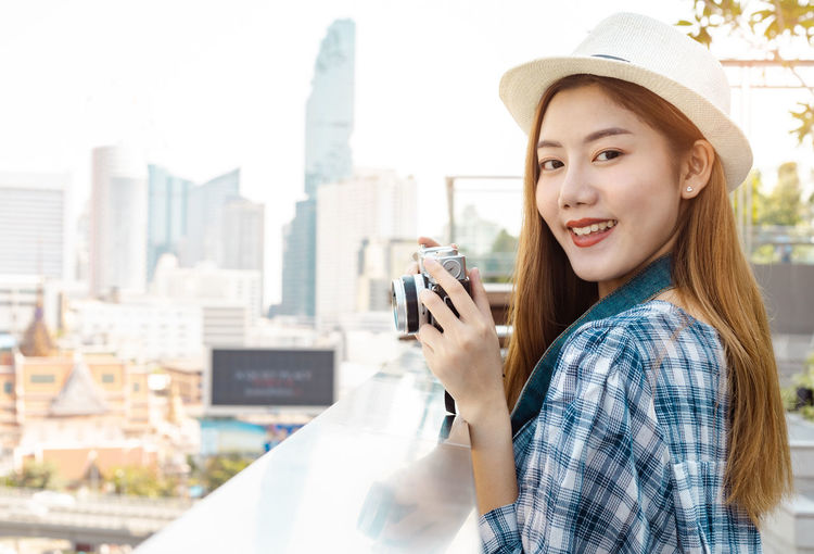 Portrait of young woman holding hat against cityscape