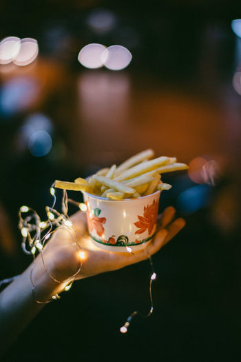 Cropped image of hand holding french fries in bowl with lighting equipment during night outdoors