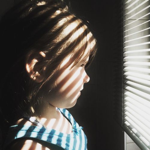 Close-up side view of girl looking through window blinds at home