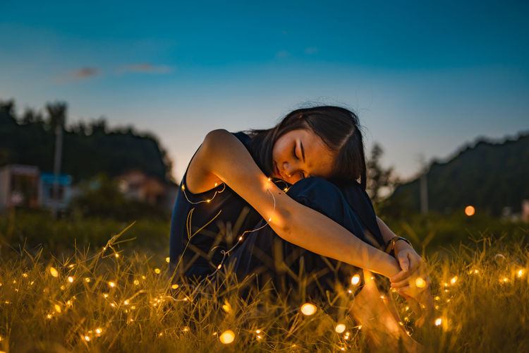 Woman with illuminated string light sitting on grass during sunset