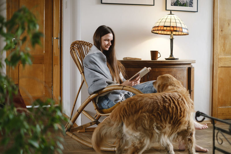 Young woman reading book with dog golden retriever, sitting on rocking chair. cozy home atmosphere