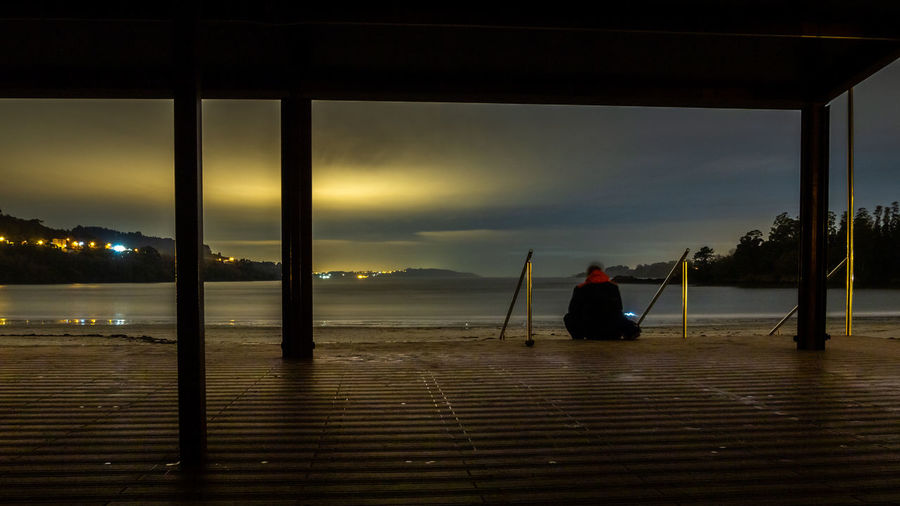 Rear view of silhouette person sitting on sea against sky during sunset