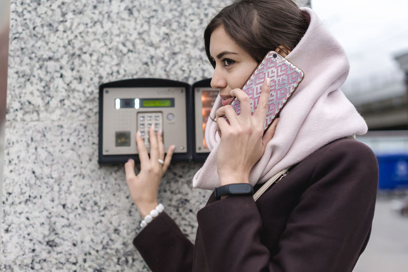 Young woman touching keypad of intercom while talking on phone