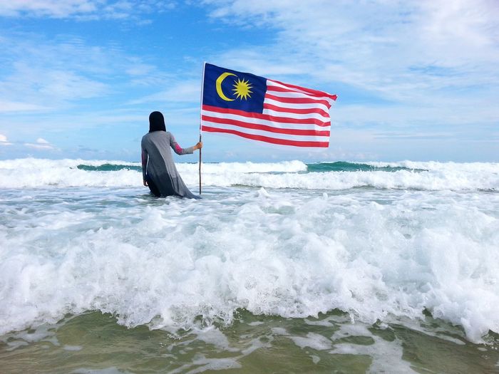 Rear view of woman standing with malaysian flag in sea against sky