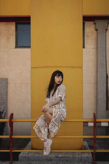 Full body of young self esteem ethnic female with long dark hair in stylish outfit sitting on metal railing near modern building and looking away