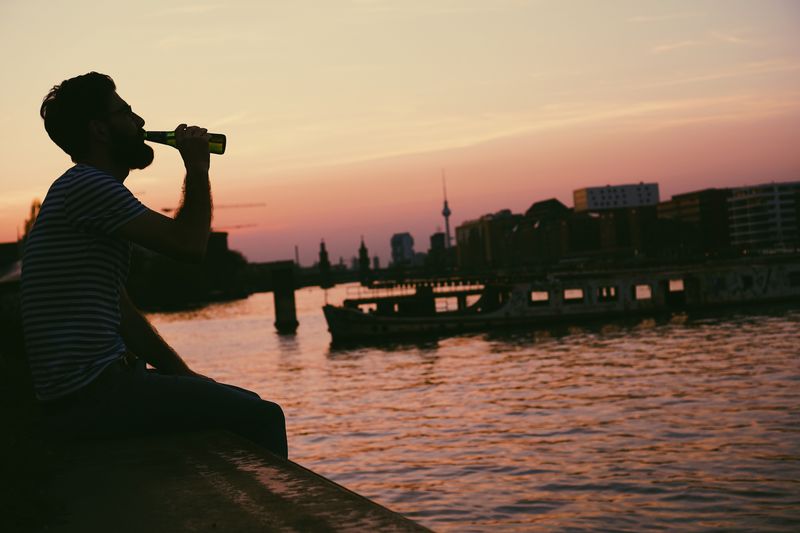Man photographing by river against sky during sunset