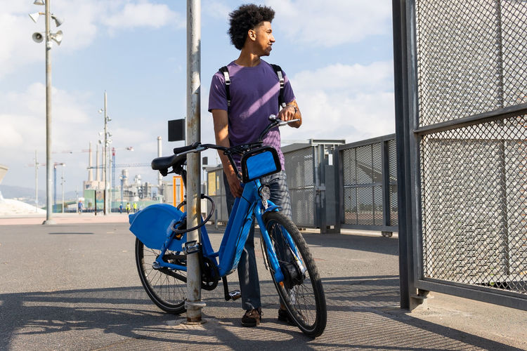 Young curly haired african american male with backpack standing near pillar with bicycle and looking away with interest