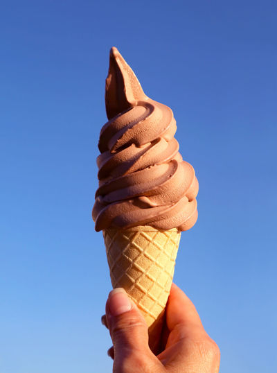 Hand holding a chocolate soft serve ice cream cone against sunny blue sky