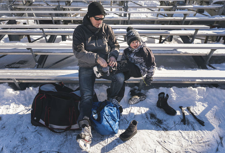 High angle view of father tying son's shoelace while sitting on bench during winter