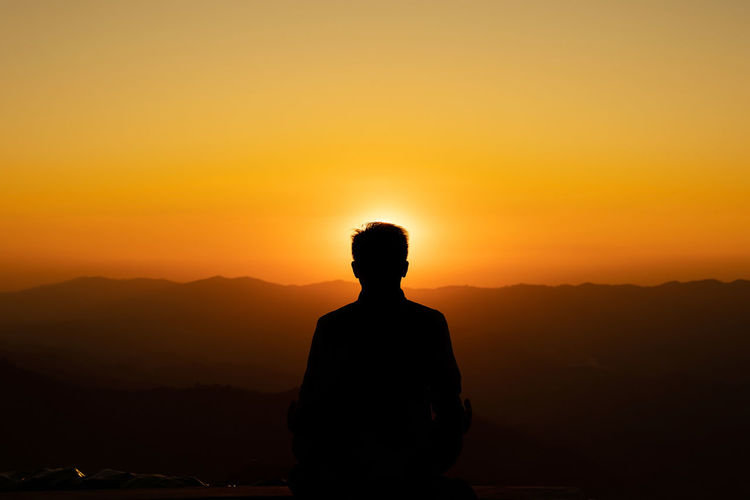Silhouette man meditating while sitting against orange sky during sunset